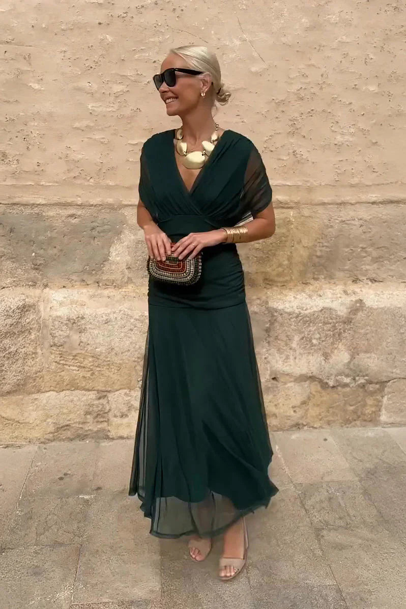 Woman wearing deep emerald V-neck maxi dress with sheer sleeves and ruched waist detail against textured wall