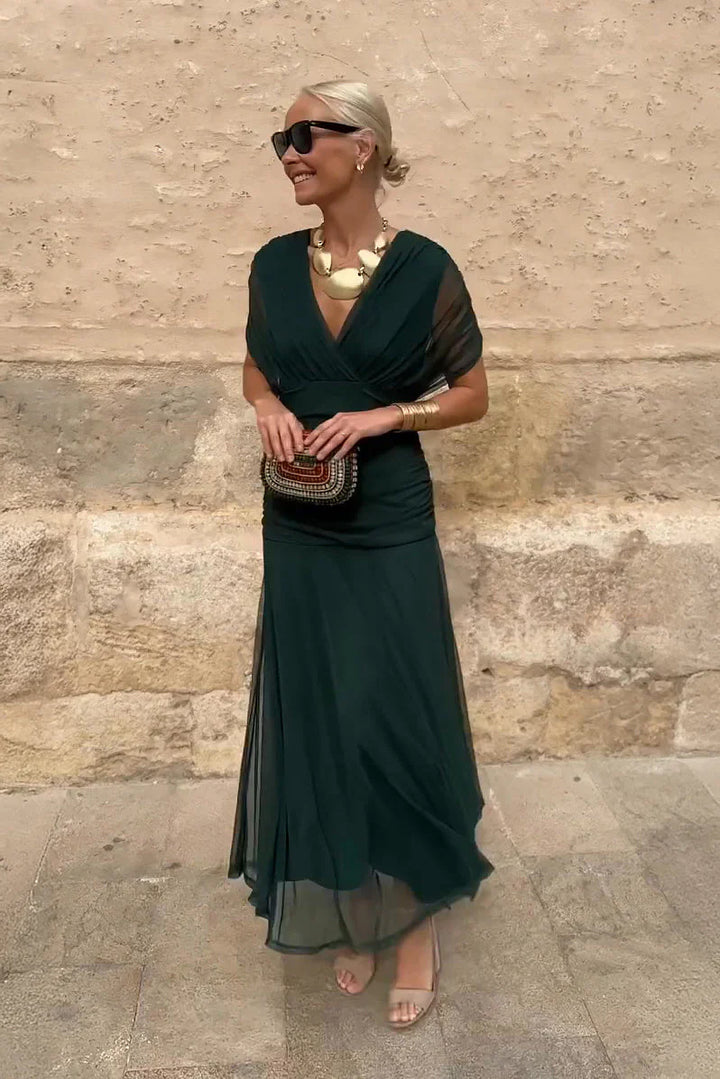 Woman wearing deep emerald V-neck maxi dress with sheer sleeves and ruched waist detail against textured wall