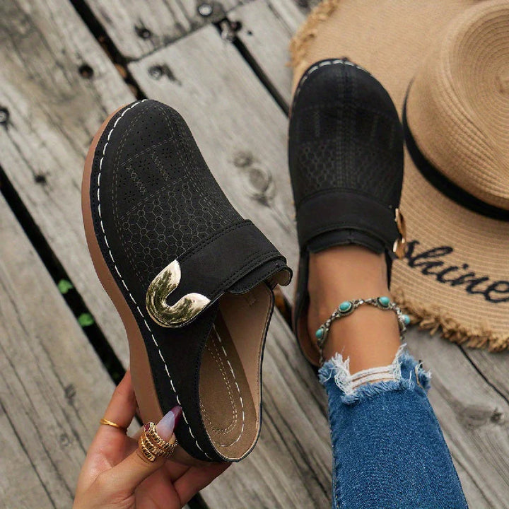 Women's slip-on mules with textured black upper and decorative gold buckle detail on wooden deck