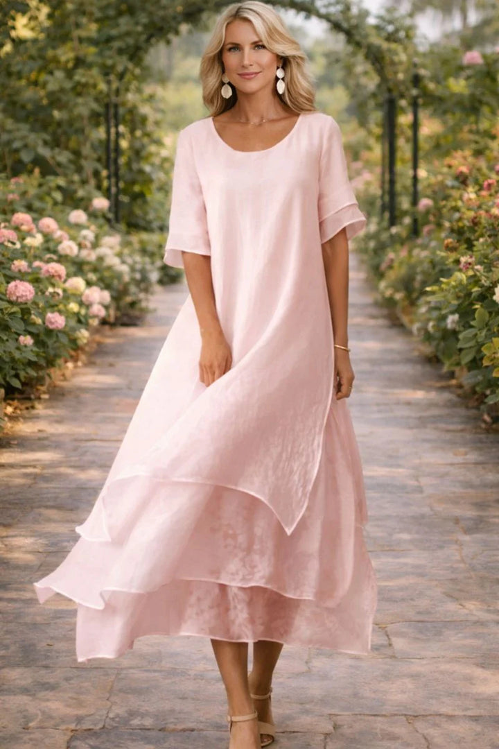 Layered midi dress in blush pink with scoop neckline and tiered hem, worn outdoors in summer garden setting